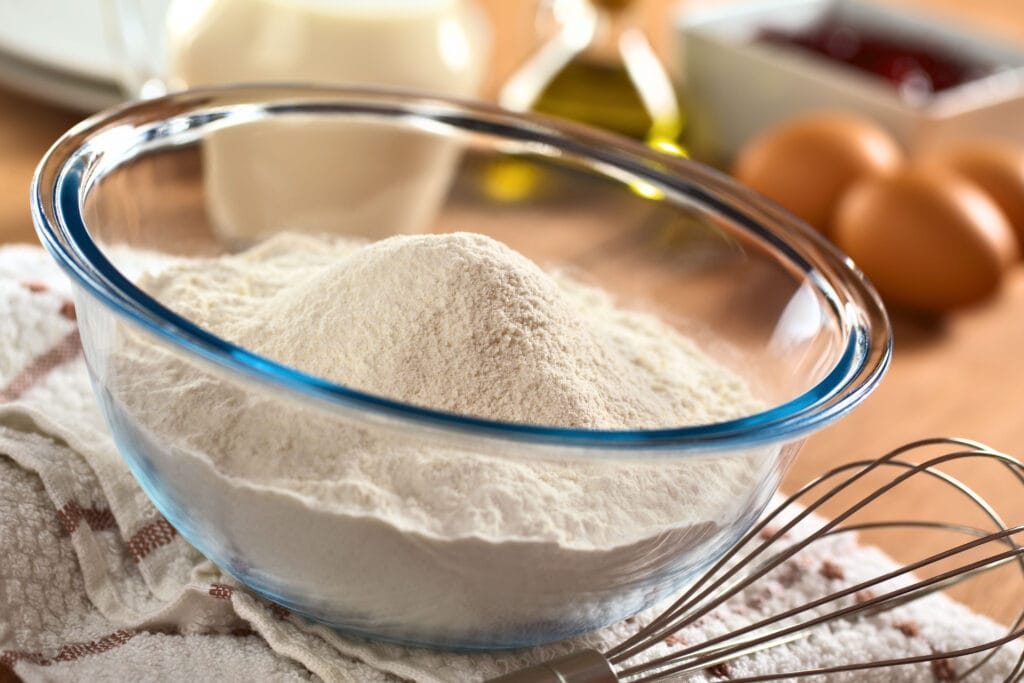 Glass Bowl of Flour Sitting on a Dish Towel with a Wire Whisk