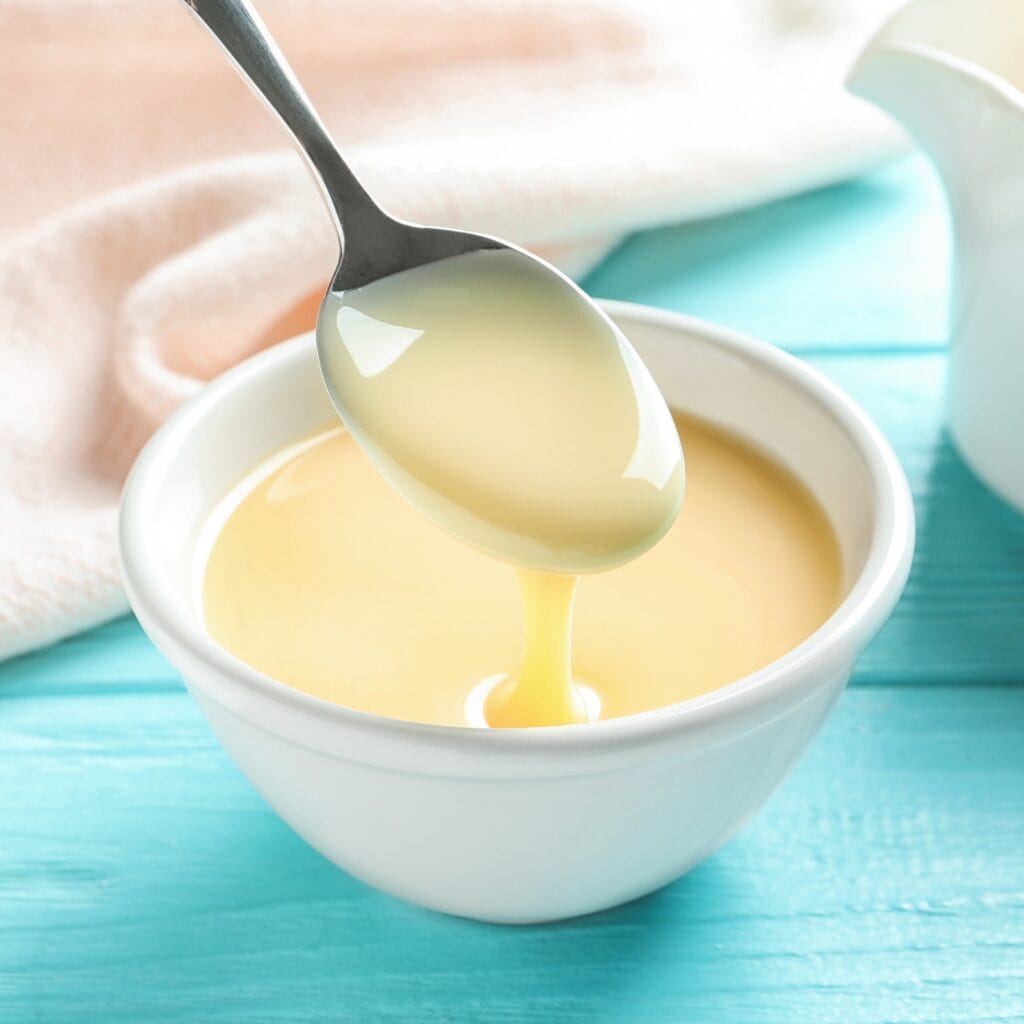 The Best Sweetened Condensed Milk Substitutes featuring Sweetened Condensed Milk Dripping Off a Spoon into a Bowl of Sweetened Condensed Milk on a Turquoise Table