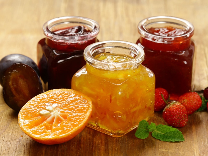 Three Small Jars of Jam and Marmalade and Various Fresh Fruits- One Jar of Orange Marmalade with Fresh Orange, One Jar of Plum Jam with Fresh Plum, and One Jar of Strawberry Jam with Fresh Strawberries
