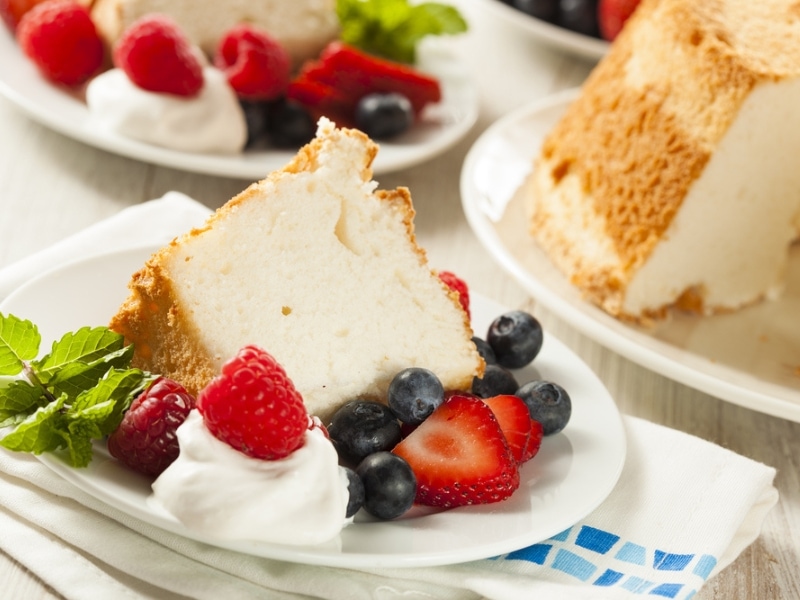 Slice of Angel's Food Cake on a Plate With Fresh Berries, Whipped Cream, and a Spring of Mint. More Similar Plates in Background.