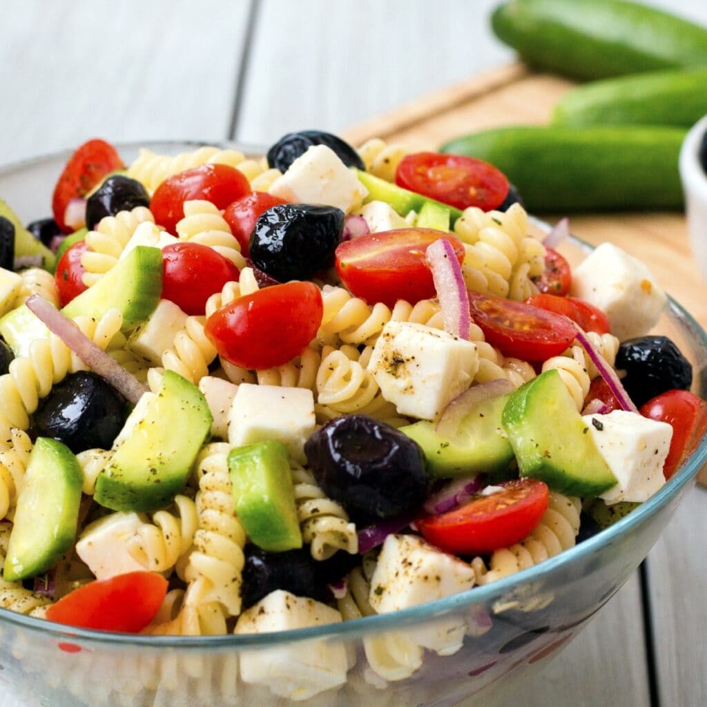 Greek Pasta Salad With Rotini, Olives, Cucumbers, Red Onion, Feta Cheese, Seasonings and Cherry Tomatoes in a Large Glass Bowl