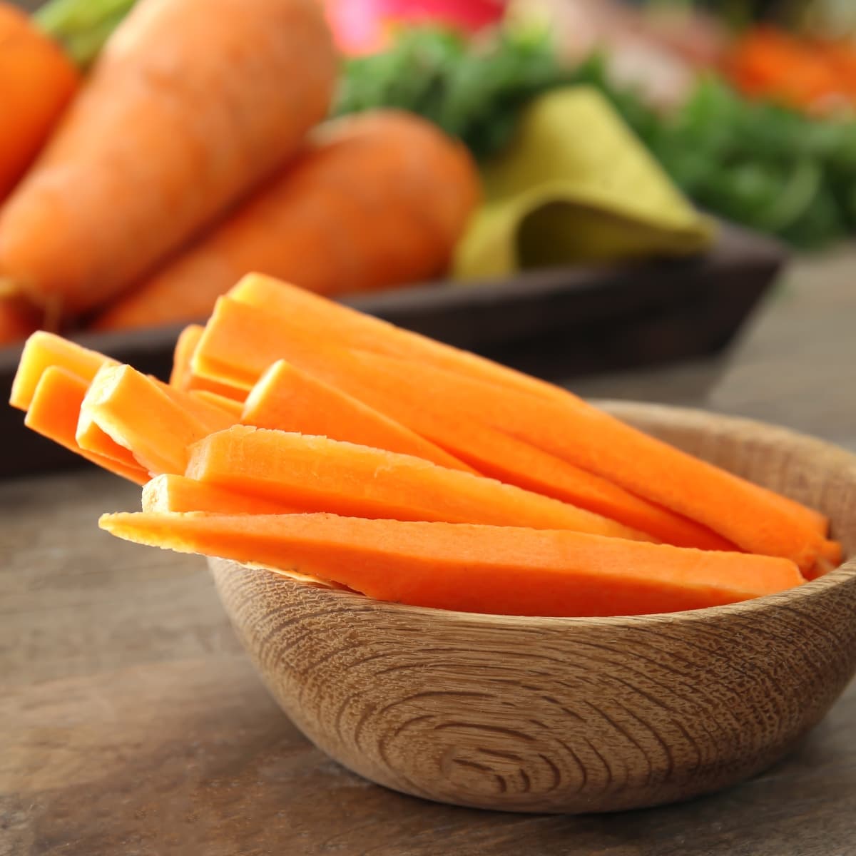 Raw Carrot Sticks in a Wooden Bowl on a Wooden Table with Whole Raw Carrots in the Background