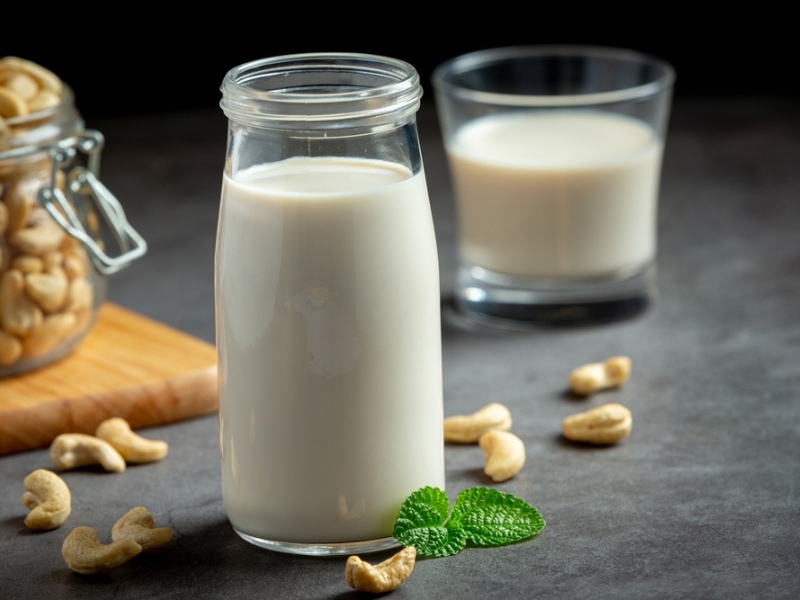 Cashew Nuts in a Glass Jar on a Wooden Cutting Board and Glass Jar of Cashew Milk in Foreground with Another Glass of Almond Milk in the Background