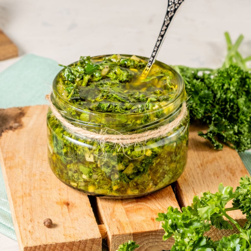 A Jar of Chimichurri Sauce on a Wooden Board with Parsley on the Side