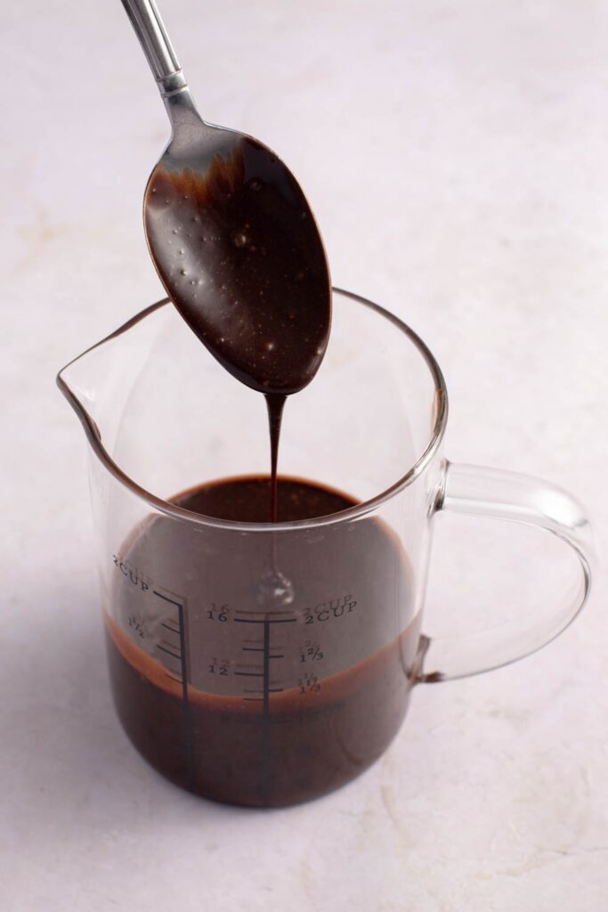 The Best Chocolate Glaze featuring Chocolate Glaze Dripping from a Spoon into a Glass Measuring Cup