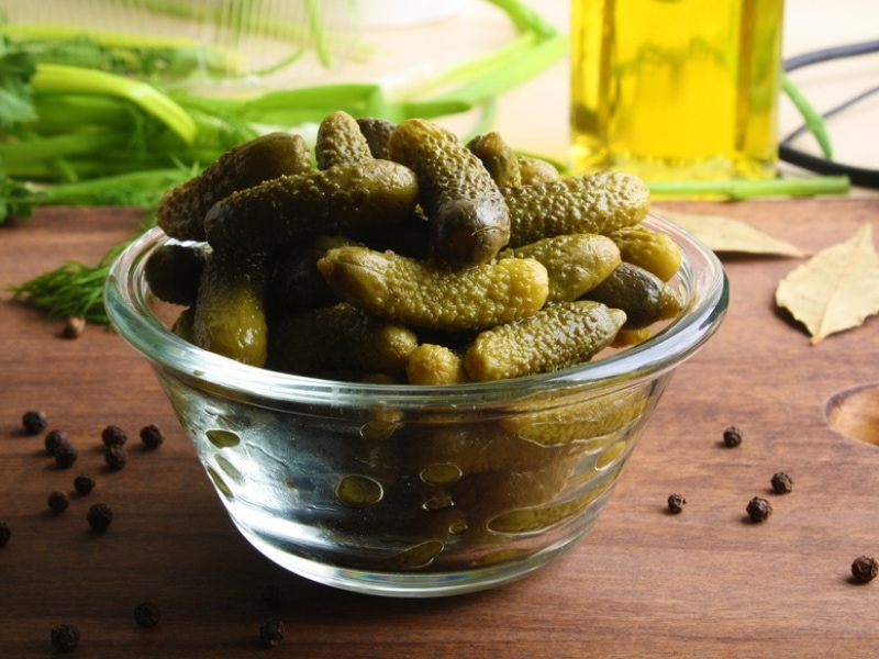Cornichons in a Clear Glass Bowl with Olive Oil, Herbs, and Black Peppercorns in the Background