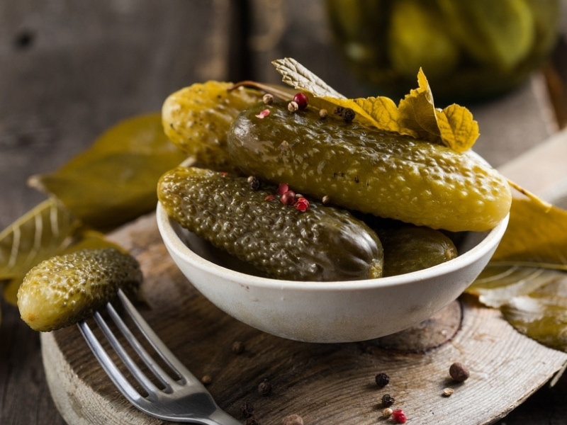German Pickles With Peppercorns in a White Bowl on a Round Cutting Board and a Fork-Speared Pickle on the Side