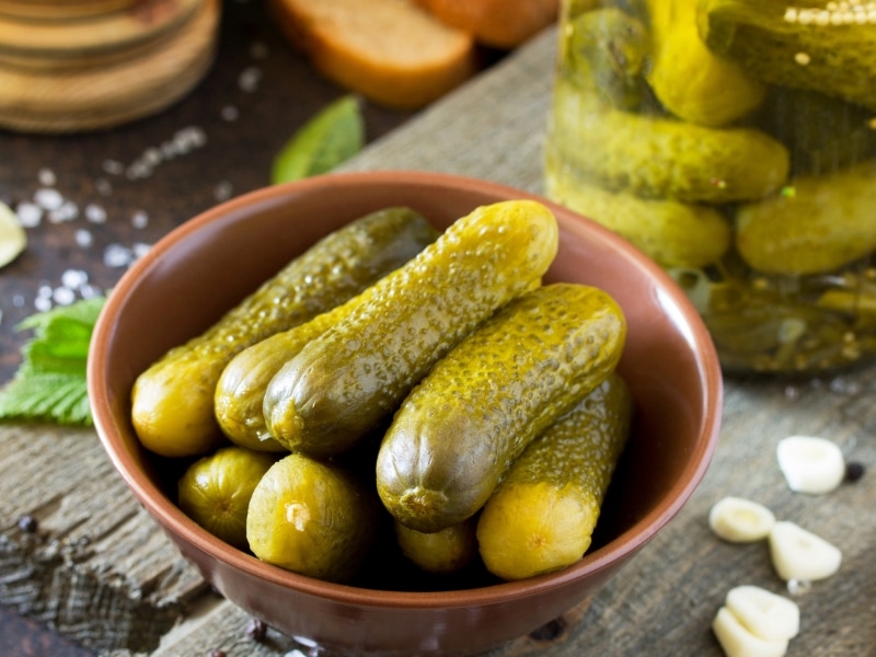 Marinated Gherkins in a Brown Bowl on a Wooden Table with More Gherkins in the Back and Cloves of Garlic
