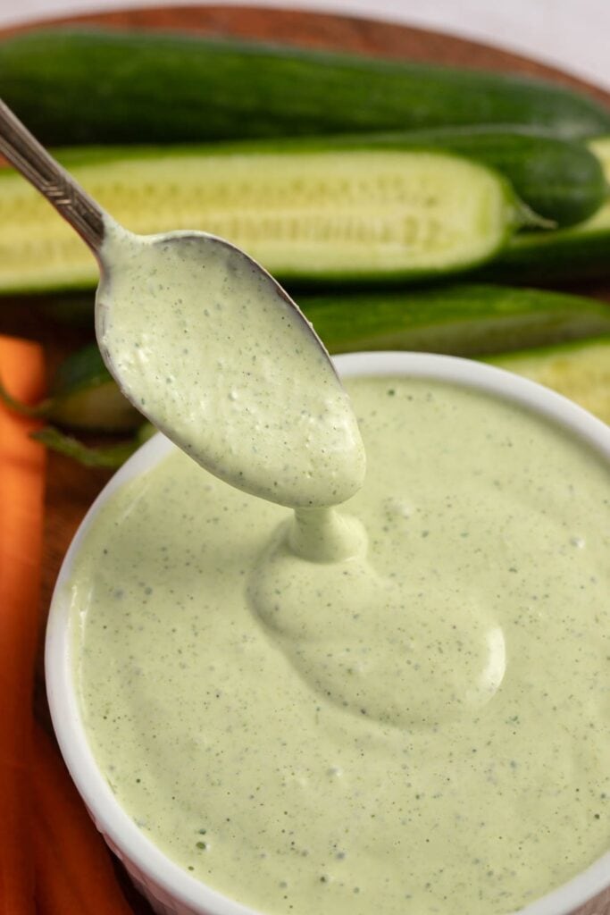 The Best Green Goddess Dressing featuring Homemade Green Goddess Dressing Dripping from Spoon Into Bowl with Cucumber and Carrots