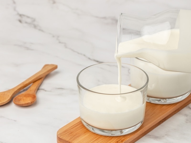 Pitcher Pouring Lactose-Free Milk Into a Glass on a Wooden Plank with Wooden Spoons to the Side