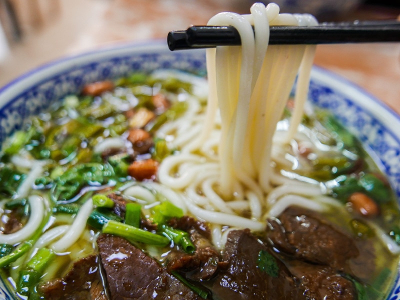 Bowl of Lamian Noodles in Broth with Chunks of Beef and Veggies, And Lamian Noodles Being Taken From Bowl with Chopsticks