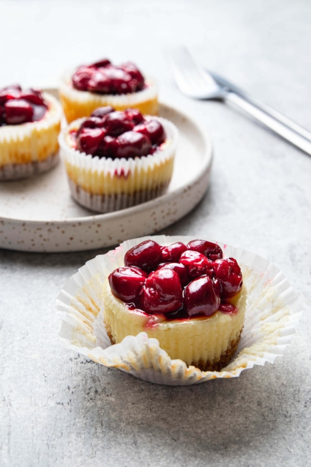 3 Mini Cheesecakes Topped With Cherries on a Plate in the Background and 1 Mini Cheesecake With Cherries in Foreground with Open Wrapper