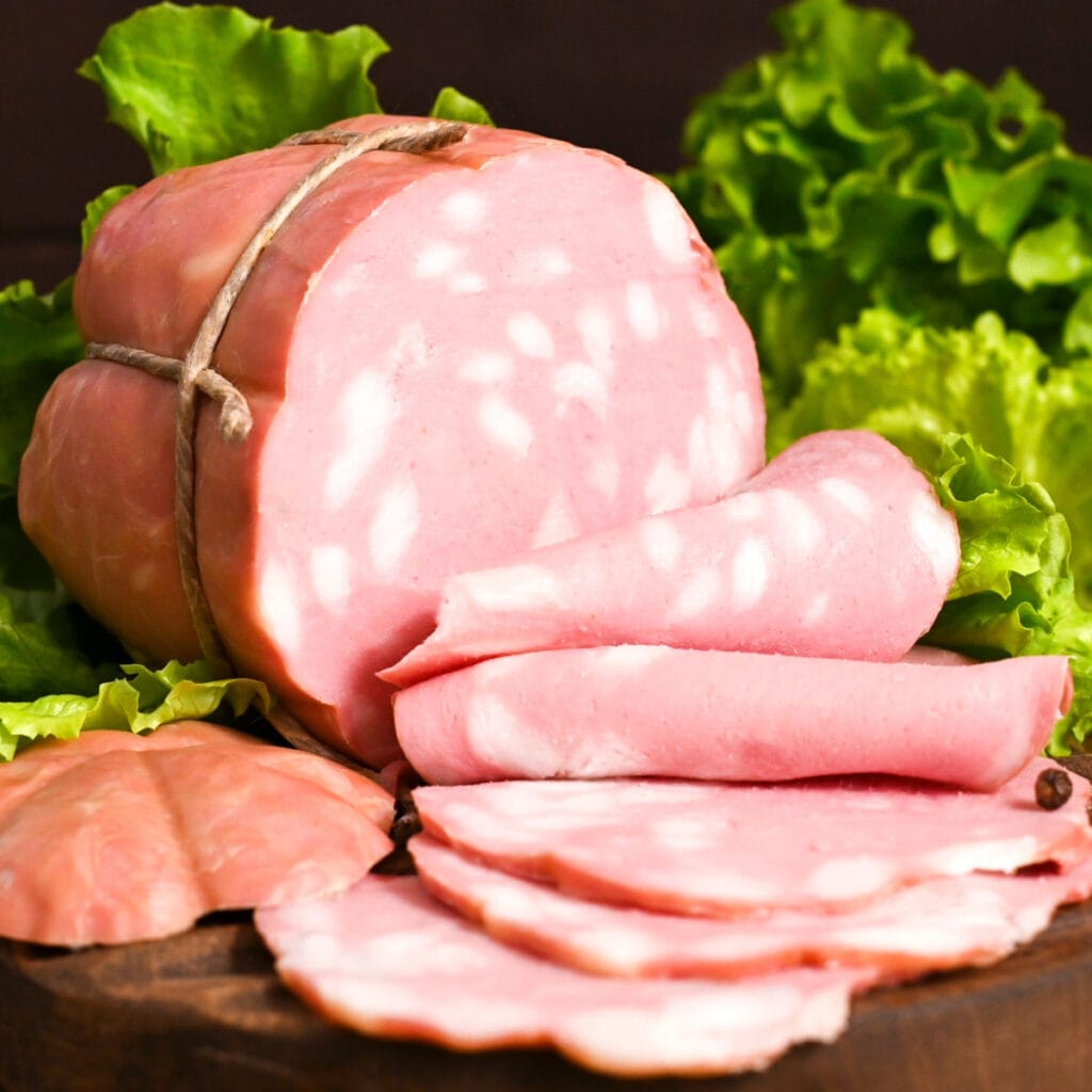 What Is Mortadella? (Everything You Need to Know) featuring Block of Mortadella With Slices on a Wooden Cutting Board, Lettuce Background