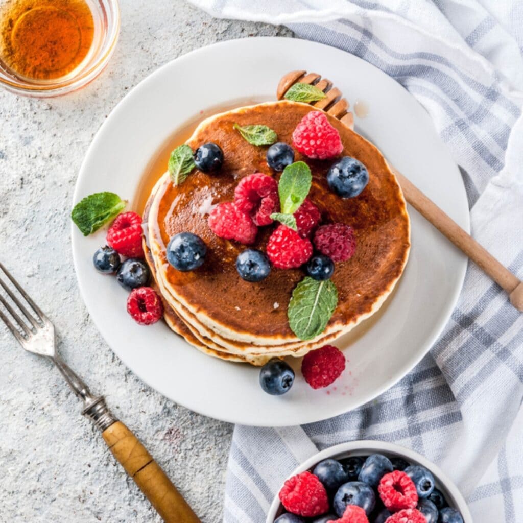 Oat Flour Pancake Top View on a Plate Topped With Fresh Berries and Dripped With Syrup and A Bowl of Berries on the Side with a Fork