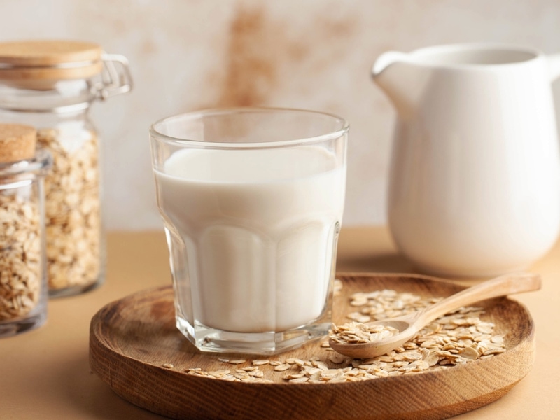 Oat Milk in a Glass on a Wooden Tray with Raw Oats and A Wooden Spoon, Pitcher of Milk and Jars of Oats in the Background