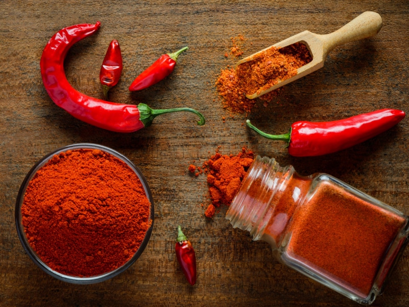 Chili Powder in a Jar and Jar of Paprika Powder on a Wooden Table Surrounded by Fresh Red Peppers