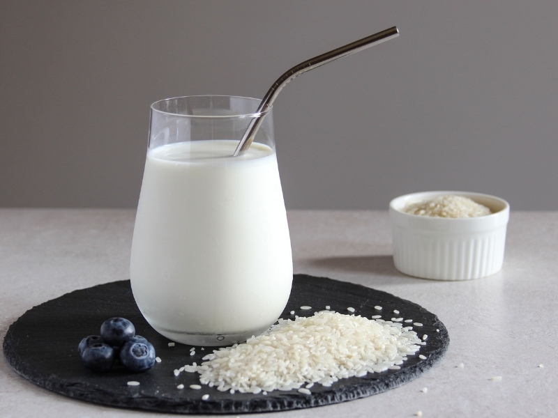 Glass of Rice Milk with a Metal Straw on a Slate Board with Rice Grains and Blueberries, Bowl of Rice in the Background