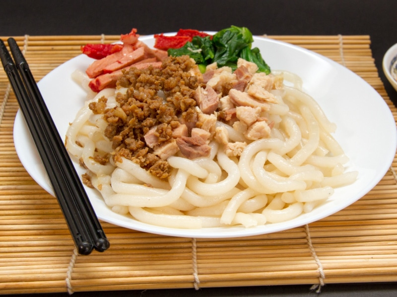 Silver Needle Noodles with Beef, Pork, and Chicken and Sauteed Greens on a Plate on a Bamboo Mat with Chopsticks