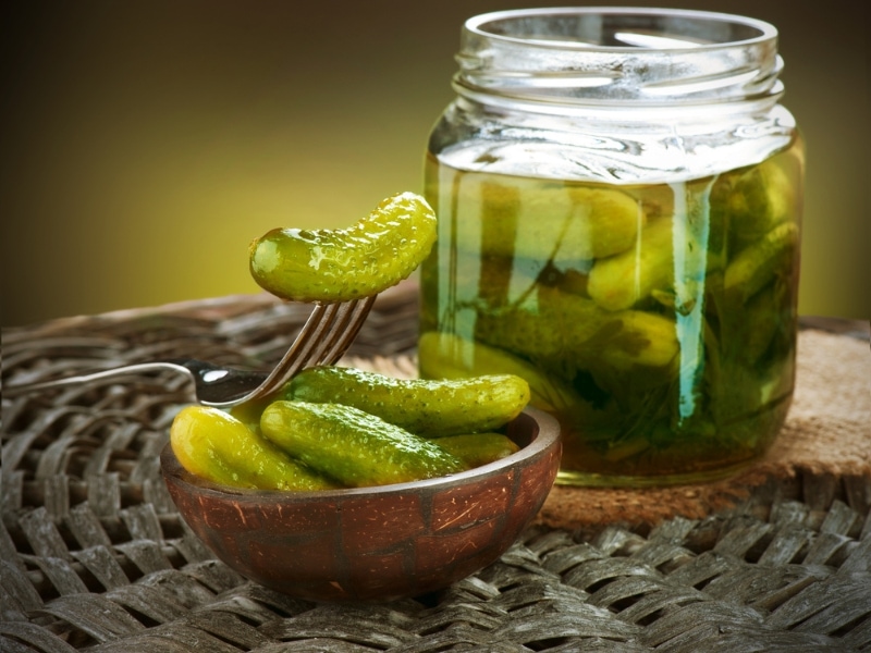 Marinated Sour Pickles in a Jar next to More Sour Pickles in a Wooden Bowl with One Fork-Speared Pickle