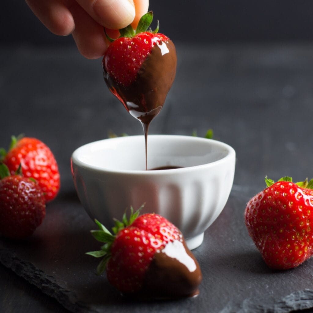 Strawberry Being Dipped into Small Bowl of Chocolate with Fresh Strawberries Around the Bowl and One Chocolate-Dipped Strawberry in Front
