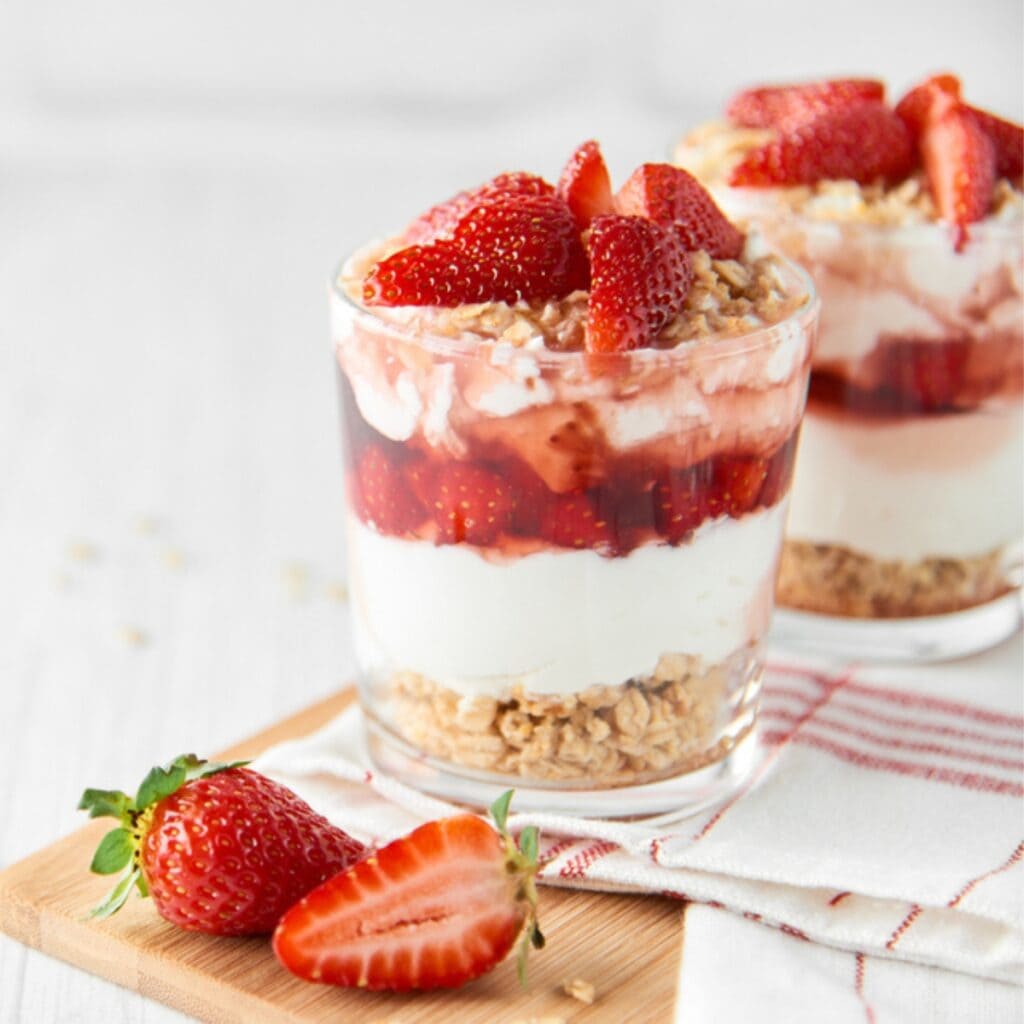 Homemade Strawberry Parfait With Layers of Yogurt, Granola, and Strawberries on a White and Red Checked Cloth on A Cutting Board with Fresh Strawberries on the Side