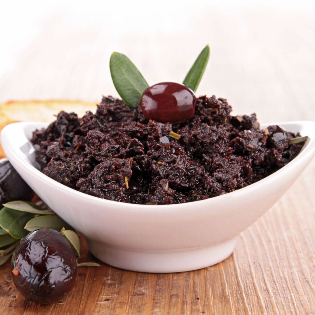 Bowl of Black Olive Tapenade Garnished with a Black Olive on a Wooden Table and More Black Olives on the Side