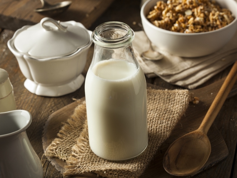 Glass Bottle of White Whole Milk on a Burlap Cloth with a Bowl of Granola, a Wooden Spoon, and a Ceramic Bowl with a Lid in the background, on a Wooden Table