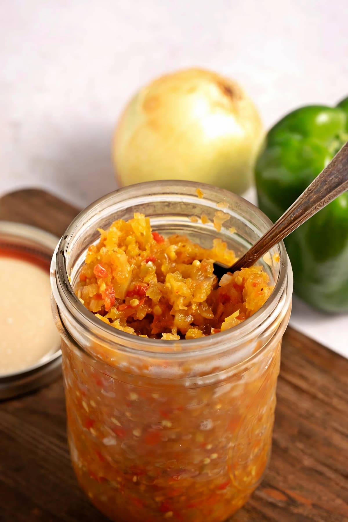 A Mason Jar of Sweet and Tangy Homemade Green Tomato Relish on a Wooden Cutting Board with Peppers and Onions in the Background