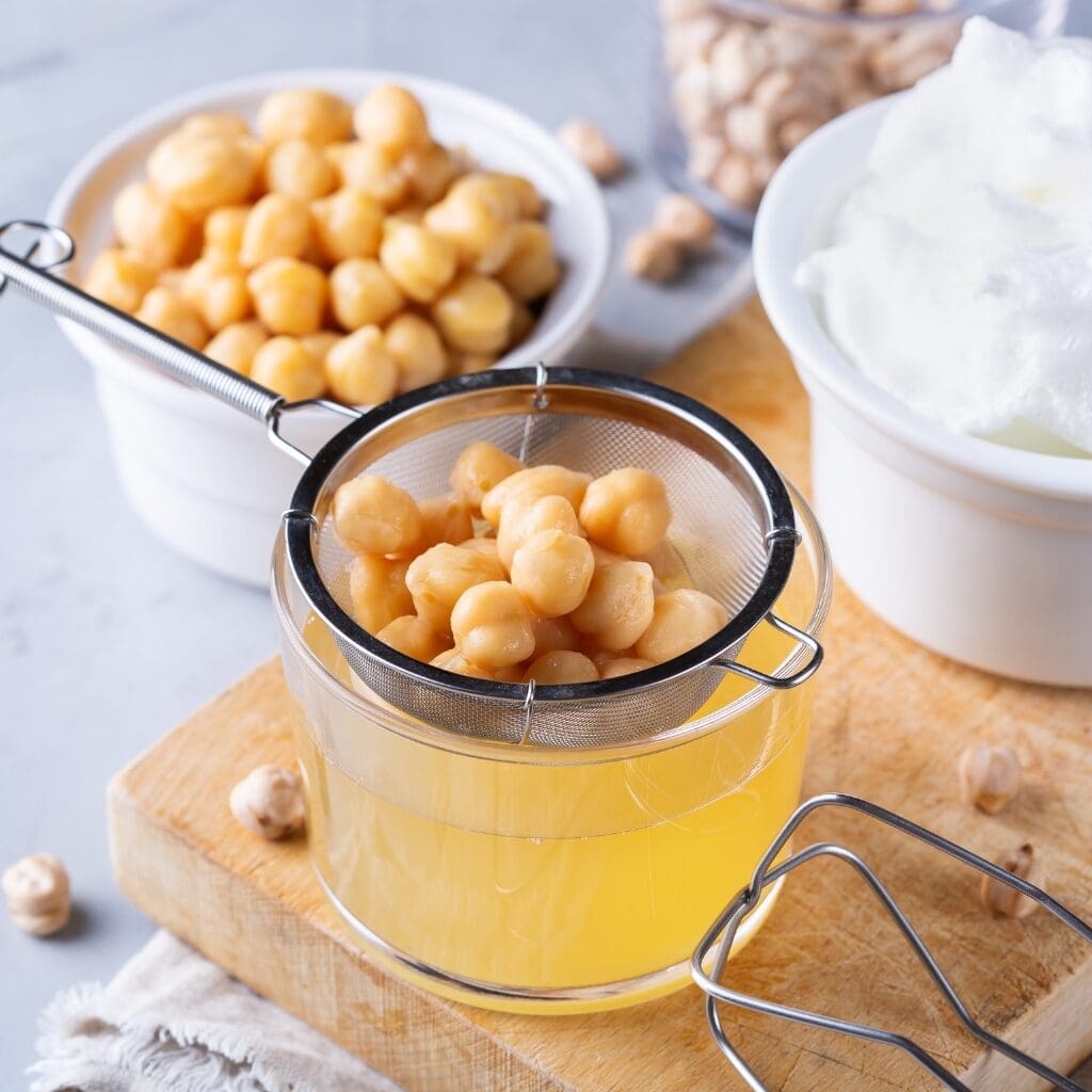 Chickpeas in a Strainer, Draining into a Glass Jar of Aquafaba with More Chickpeas in a Bowl in the Background