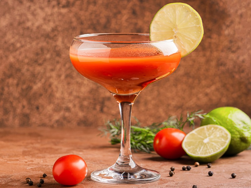Tomato Juice Bloody Maria Garnished with Lime in a Martini Glass with Tomatoes, Limes, Peppercorns, and Rosemary in the Background