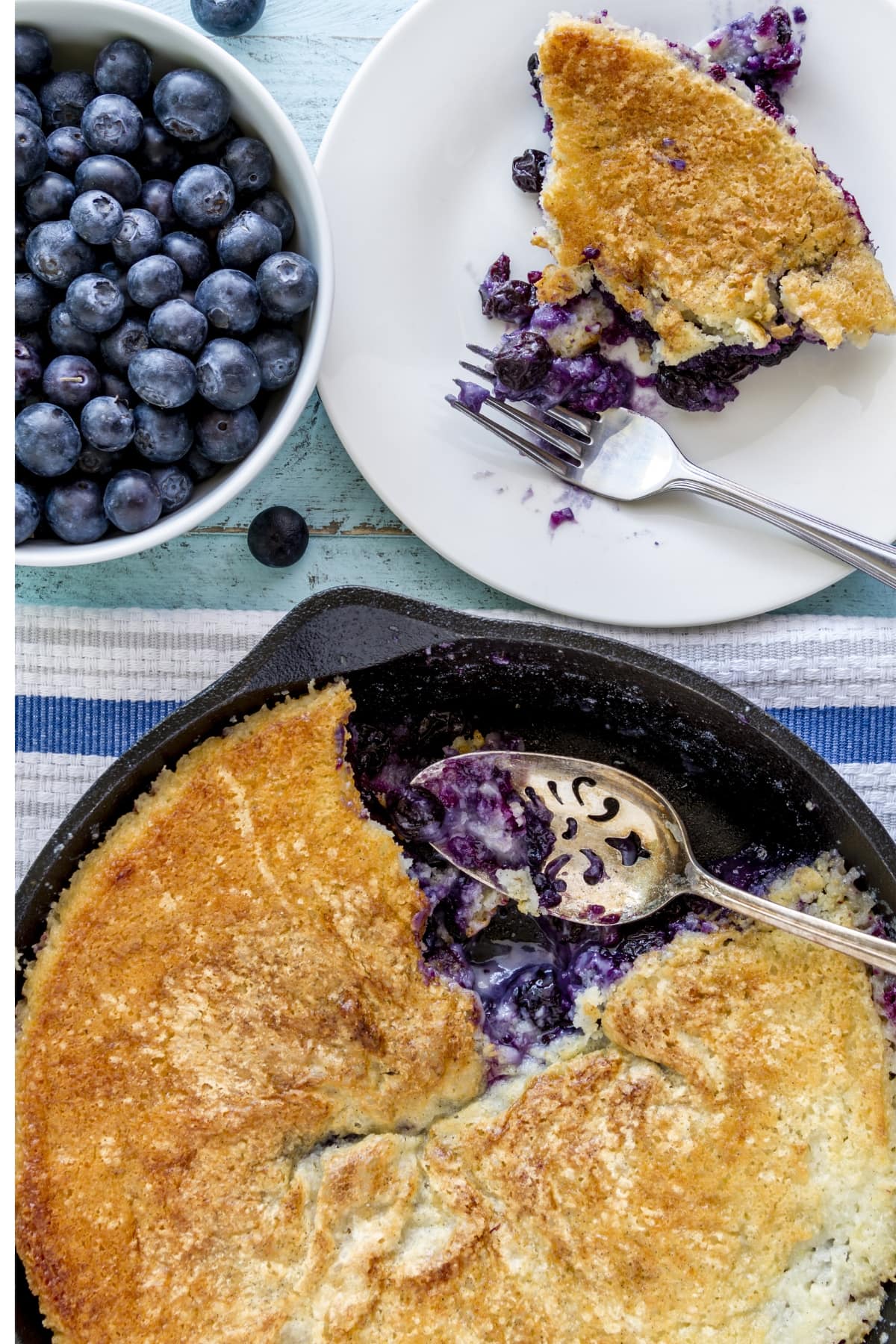 Cobbler vs. Pie (What's the Difference?) featuring Blueberry Cobbler in a Cast Iron Pan and A Slice on a Small White Plate with a Fork and A Side of Fresh Blueberries in a Bowl