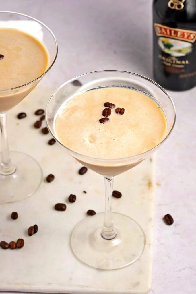 Two Martini Glasses filled with Creamy Homemade Espresso Martinis with Baileys, Garnished with Coffee Beans and a Bottle of Baileys in Background