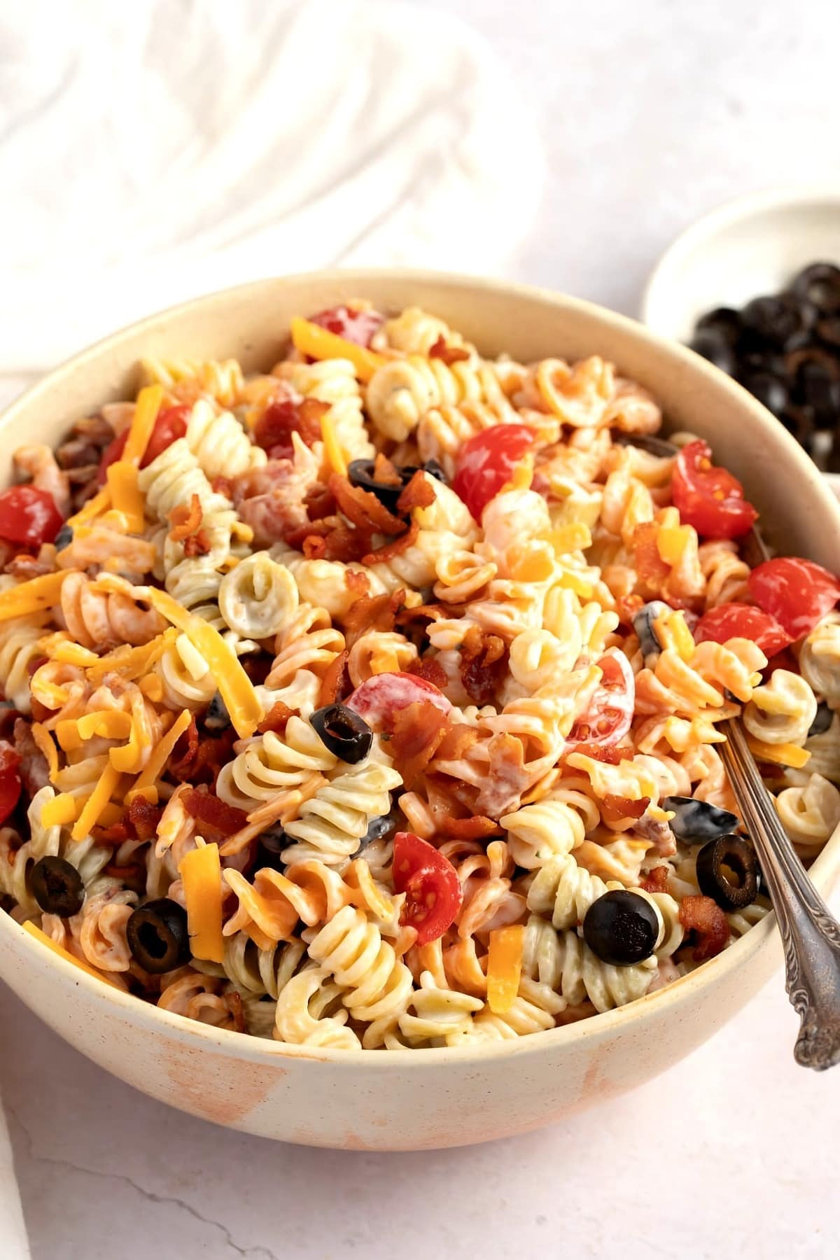 Close up view of a bowl of rotini salad with bacon, mayonnaise, milk, tomato, black olives and cheese.