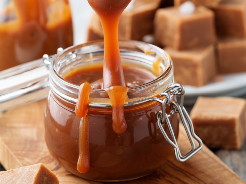 Caramel Sauce Pouring into a Jar of Caramel Sauce on a Wooden Board with Square Caramel Cadies in the Background