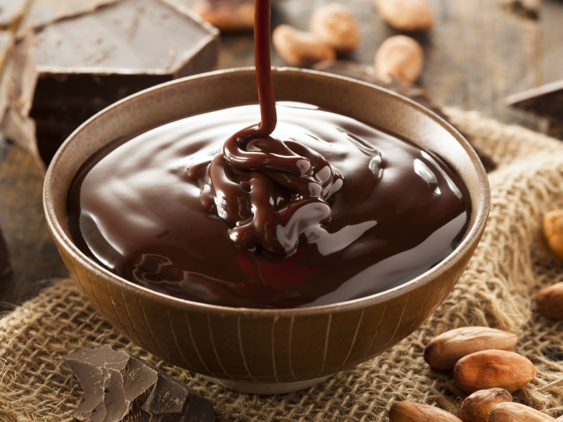 Chocolate Syrup Pouring into a Bowl of Chocolate Syrup on a Burlap Cloth with Chunks of Dark Chocolate and Nuts All Around the Bowl