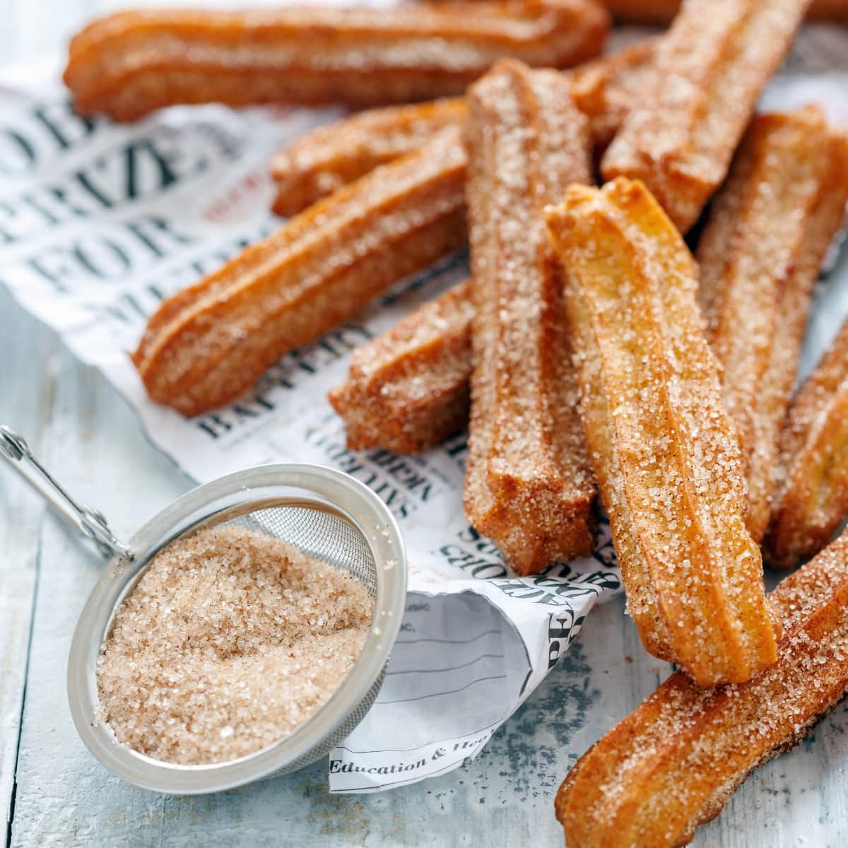 Homemade Churros Dusted With Homemade Cinnamon Sugar on a Newspaper with Cinnamon Sugar in a Dusting Sieve Beside It