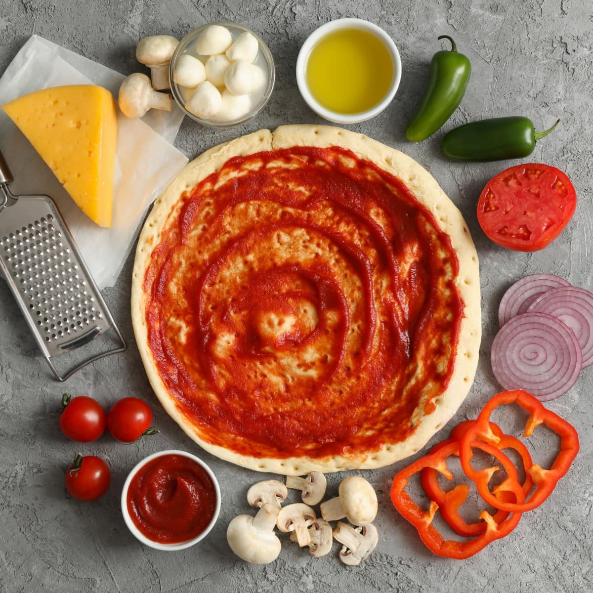 Homemade Pizza Ingredients- Homemade Crust Dough, Red Sauce, Block of Cheddar Cheese with Grater, Jalapenos, Red Onion Slices, Mushrooms, Tomatoes, Bell Peppers, Butter, and Mozzarella