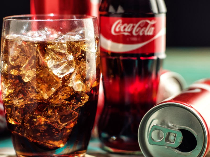 A Glass of Coca Cola over Ice with a Coca Cola Glass Bottle Behind it and an Empty Coca Cola Can Next To It