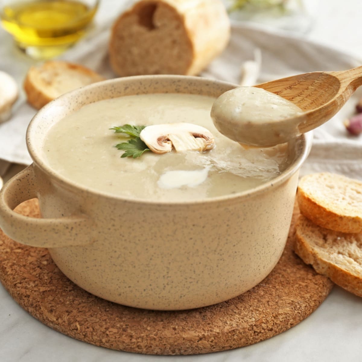 Pot of Creamy Mushroom Soup and a Wooden Spoon with Mushroom Soup on a Cork Trivet with Baguette Slices