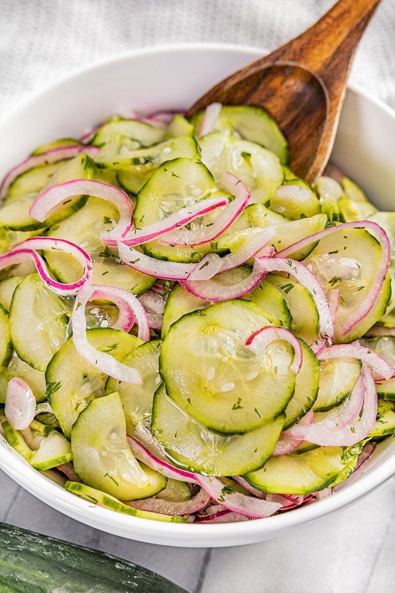 Cucumber salad with sliced onion tossed by a wooden spoon on a white bowl.