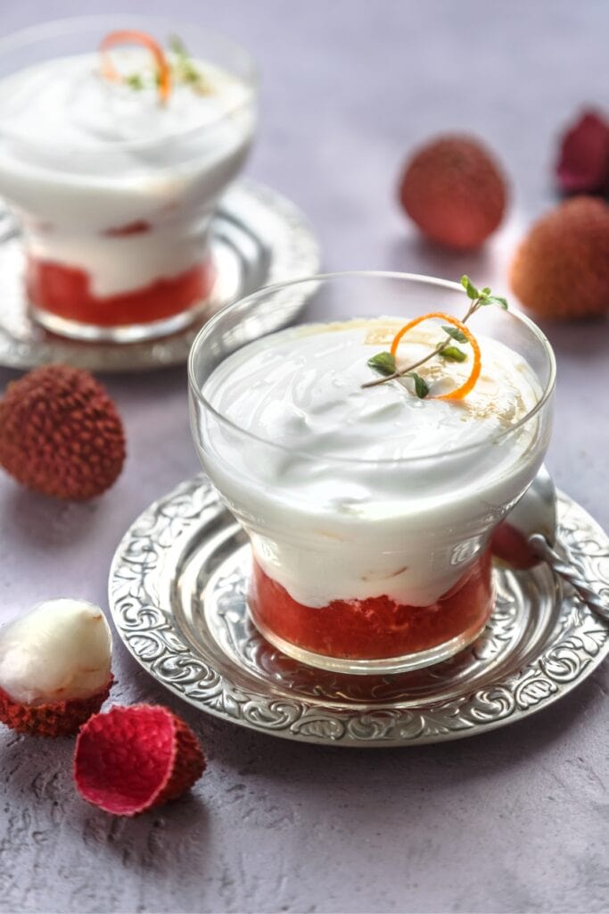 10 Easy Lychee Recipes (Fresh or Canned) featuring Glass of Lychee Dessert Topped with Cream on a Silver plate with Fresh Lychee On the Sides and In the Background