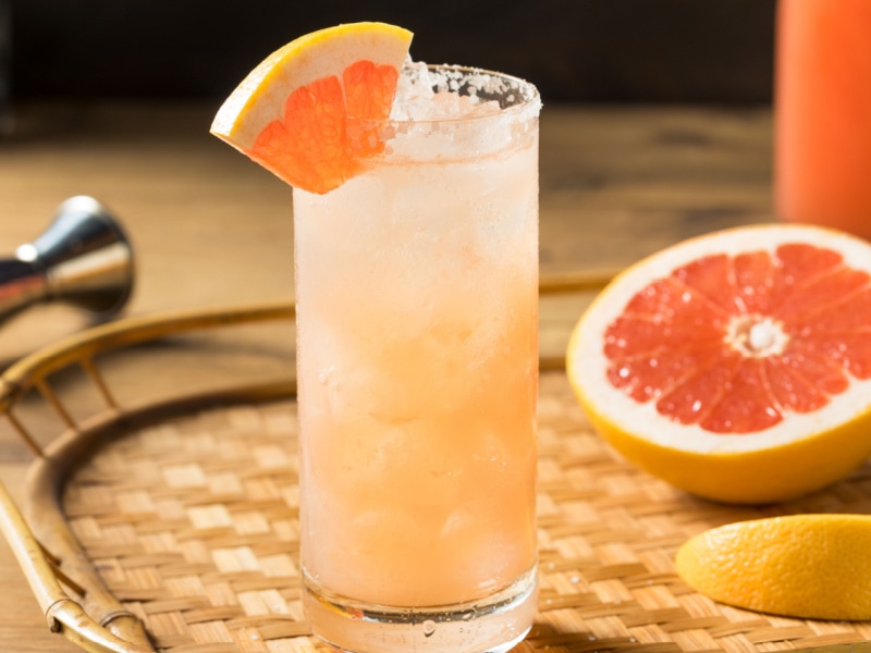Refreshing Boozy Tequila Grapefruit Paloma In a Tall Glass Over Ice with a Salted Rim Garnished with Fresh Grapefruit Slice on a Bamboo Tray with More Sliced Grapefruit Behind it