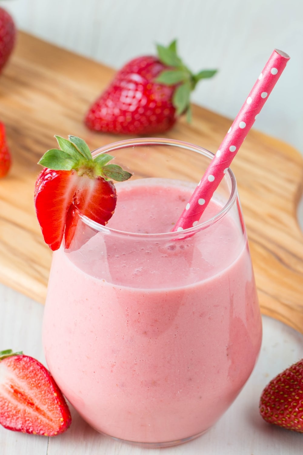 Creamy and Healthy Strawberry Smoothie in a Glass with a Pink Polka Dot Straw and a Sliced Strawberry Garnish with a Cutting Board in the Background and Fresh Strawberries Around