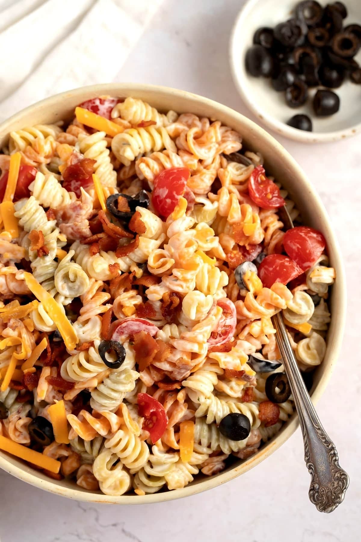Top view of a bowl of rotini salad with bacon, mayonnaise, milk, tomato, black olives and cheese.