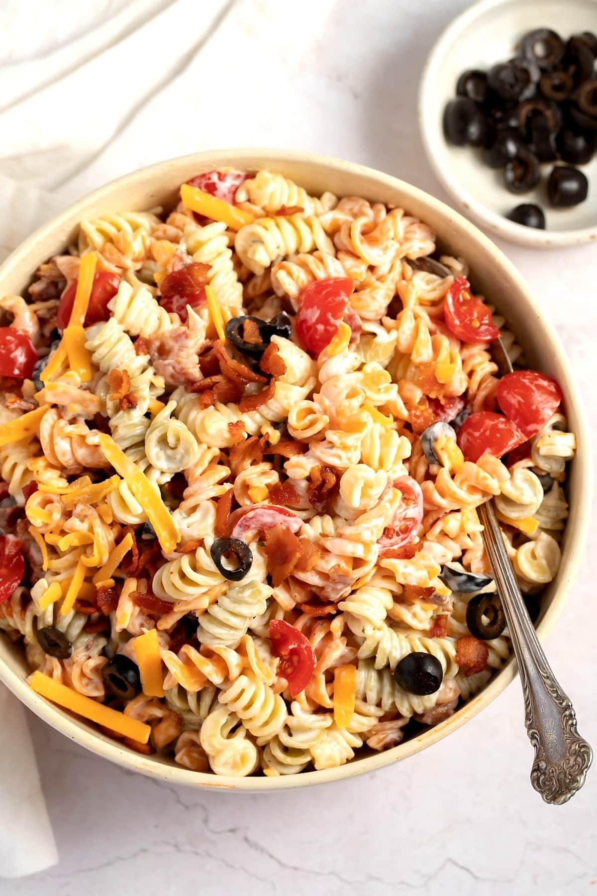 Bowl of salad made with rotini pasta, bacon, mayonnaise, garlic pepper and powder, milk, tomato, black olives and cheese