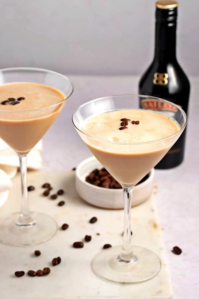 Easy Espresso Martini with Baileys featuring Two Glasses of Homemade Espresso Martinis with Baileys Garnished with Coffee Beans and a Dish of Coffee Beans in the Background with a Bottle of Baileys