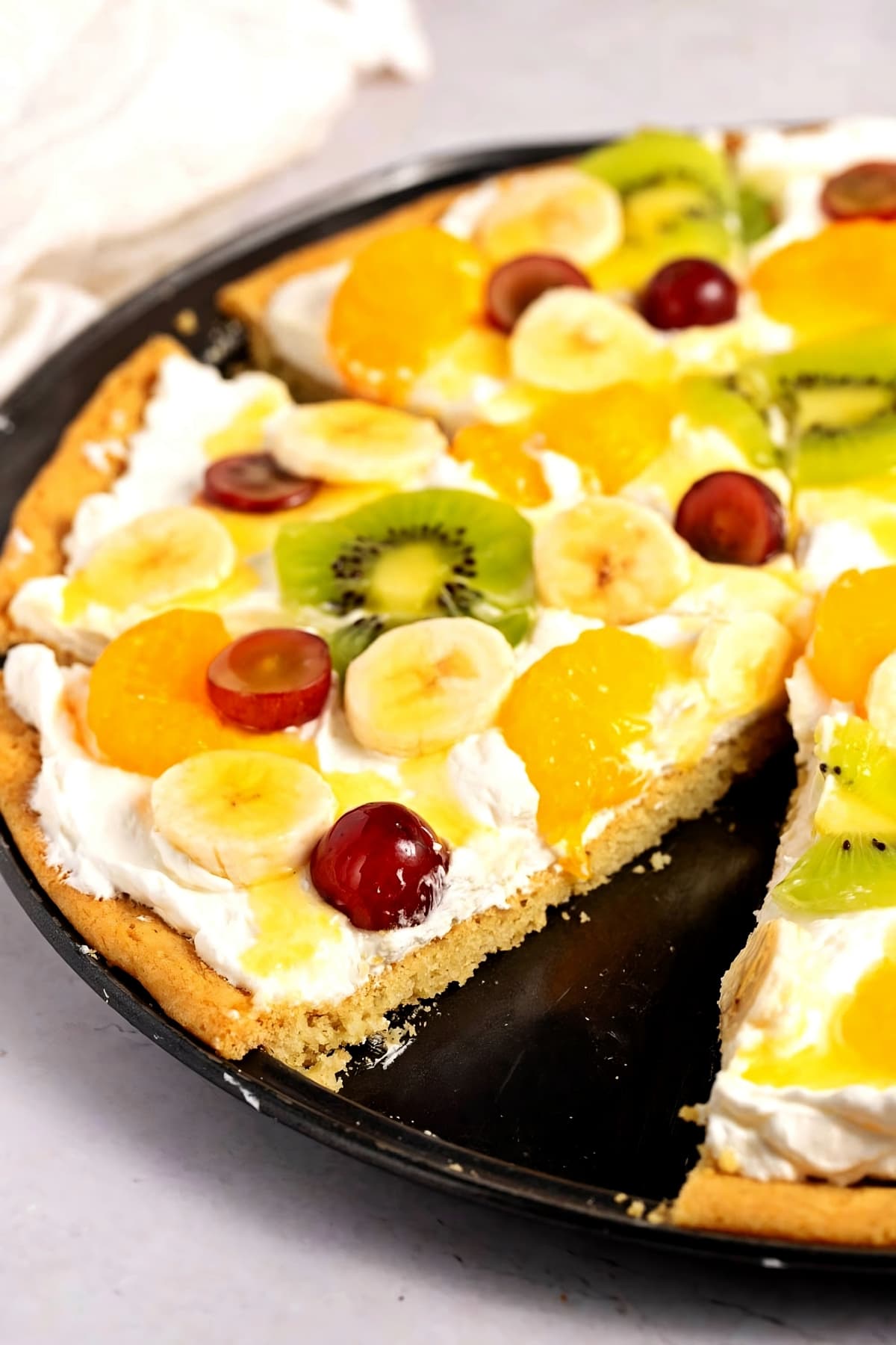 Fruit pizza topped with slices of kiwi, bananas, mandarin oranges, and grapes drizzled with lemon glaze. 