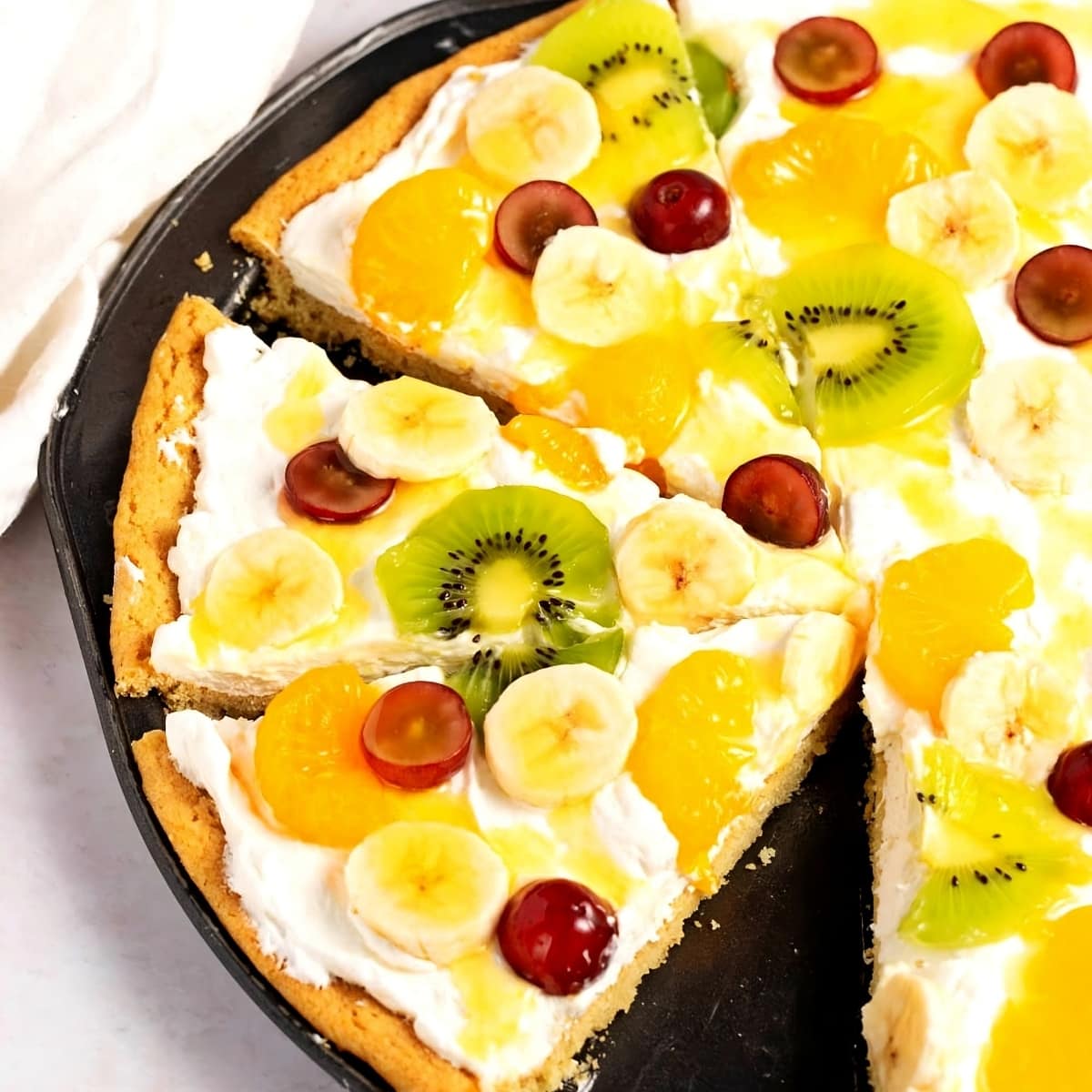 Fruit pizza in skillet pan topped with cream, kiwi, bananas, mandarin oranges, and grapes.