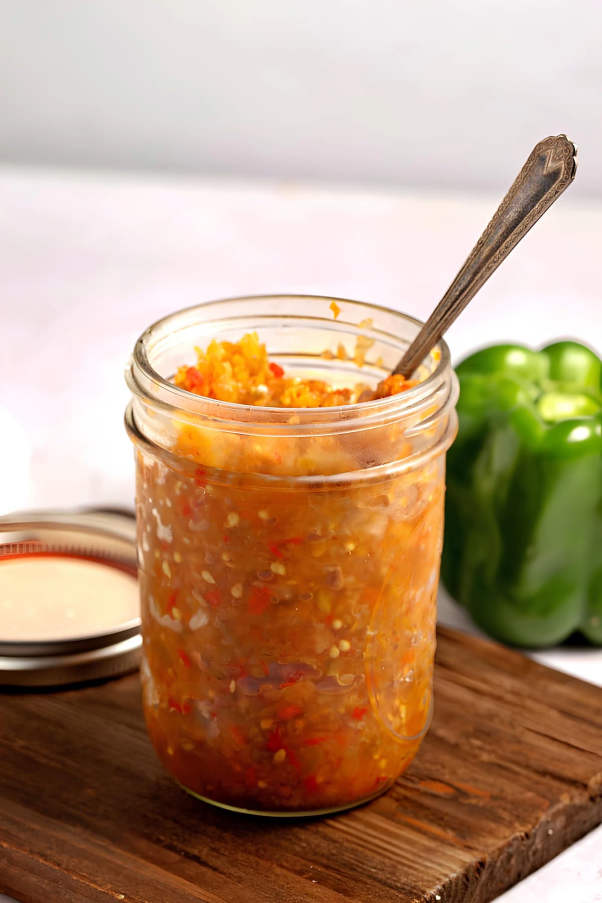 Homemade Tomato Relish in a Glass Mason Jar on a Wooden Board with Spoon in the Jar and a Pepper in the Background