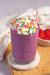 Homemade Grimace Shake Recipe with Whipped Cream and Sprinkles Featuring Raspberries in the Background
